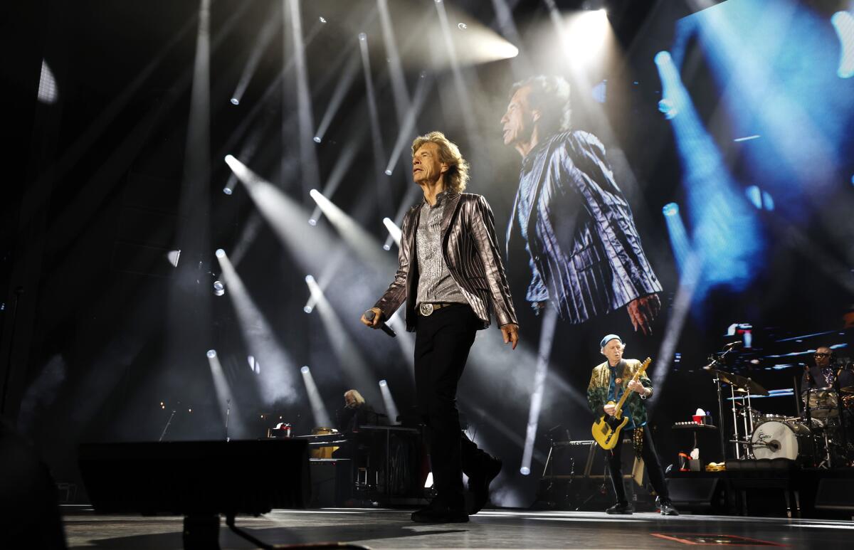 Mick Jagger walks forward in front of his bandmates onstage with an image of himself on a TV screen behind him