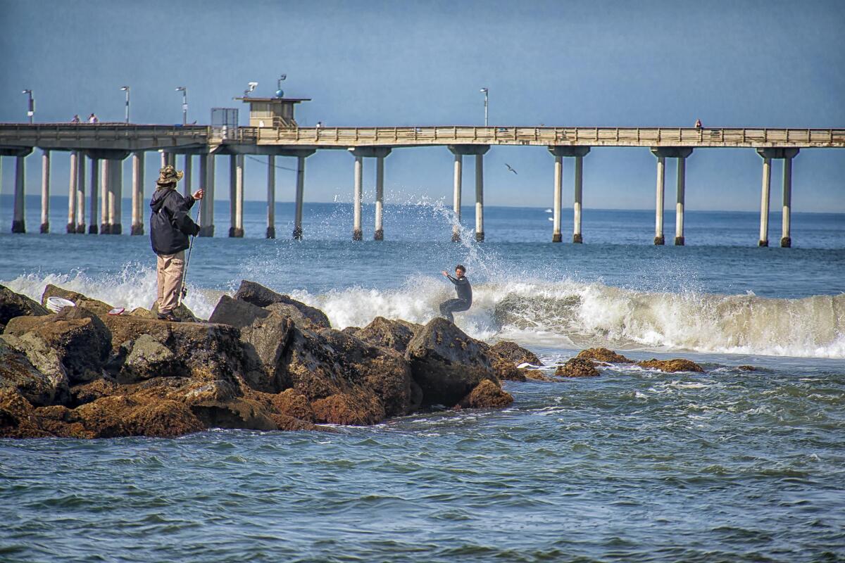 A surfer rides a wave near rocks where a fisherman stands watching with a pier in the background.