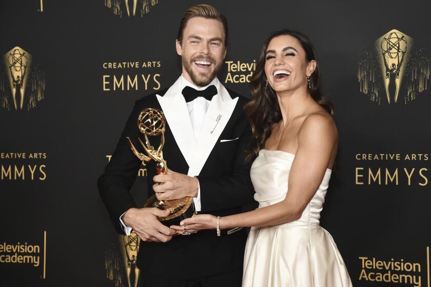 Derek Hough in a tuxedo and Hayley Erbert in a dress embrace and smile together while holding a gold statue Emmy award