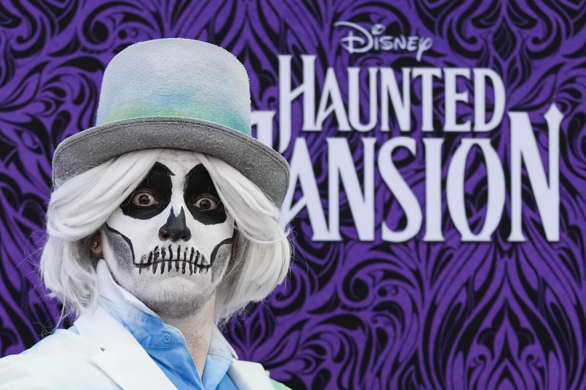 A man dressed in skeletal face paint, a white wig and a white top hat stands in front of a backdrop.