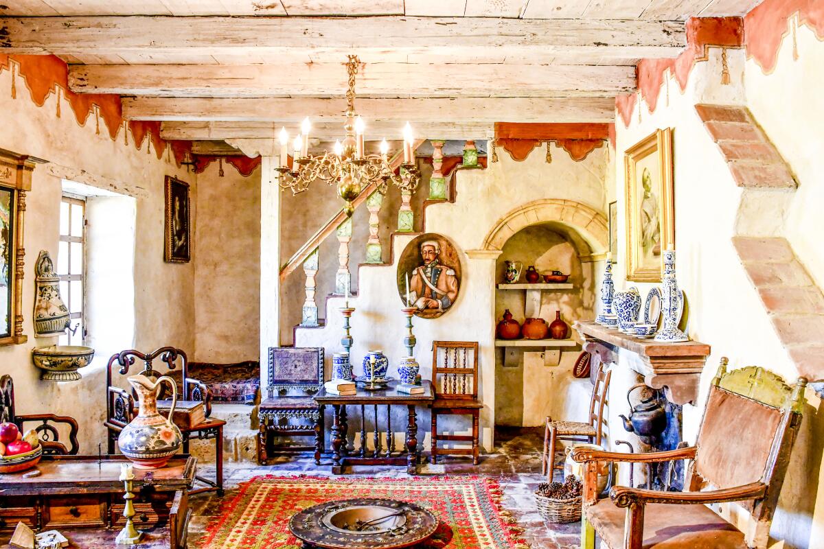 A room at Carmel Mission with ceramic items, wooden furniture and a staircase, beamed ceilings and red wall designs. 