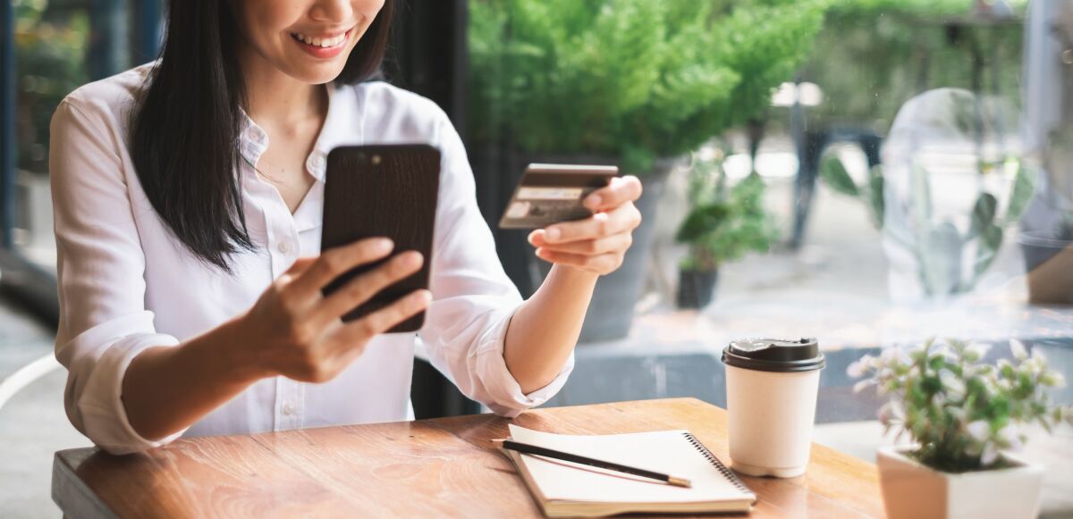 A woman at a cafe holds a cellphone in one hand and a credit card in another hand.
