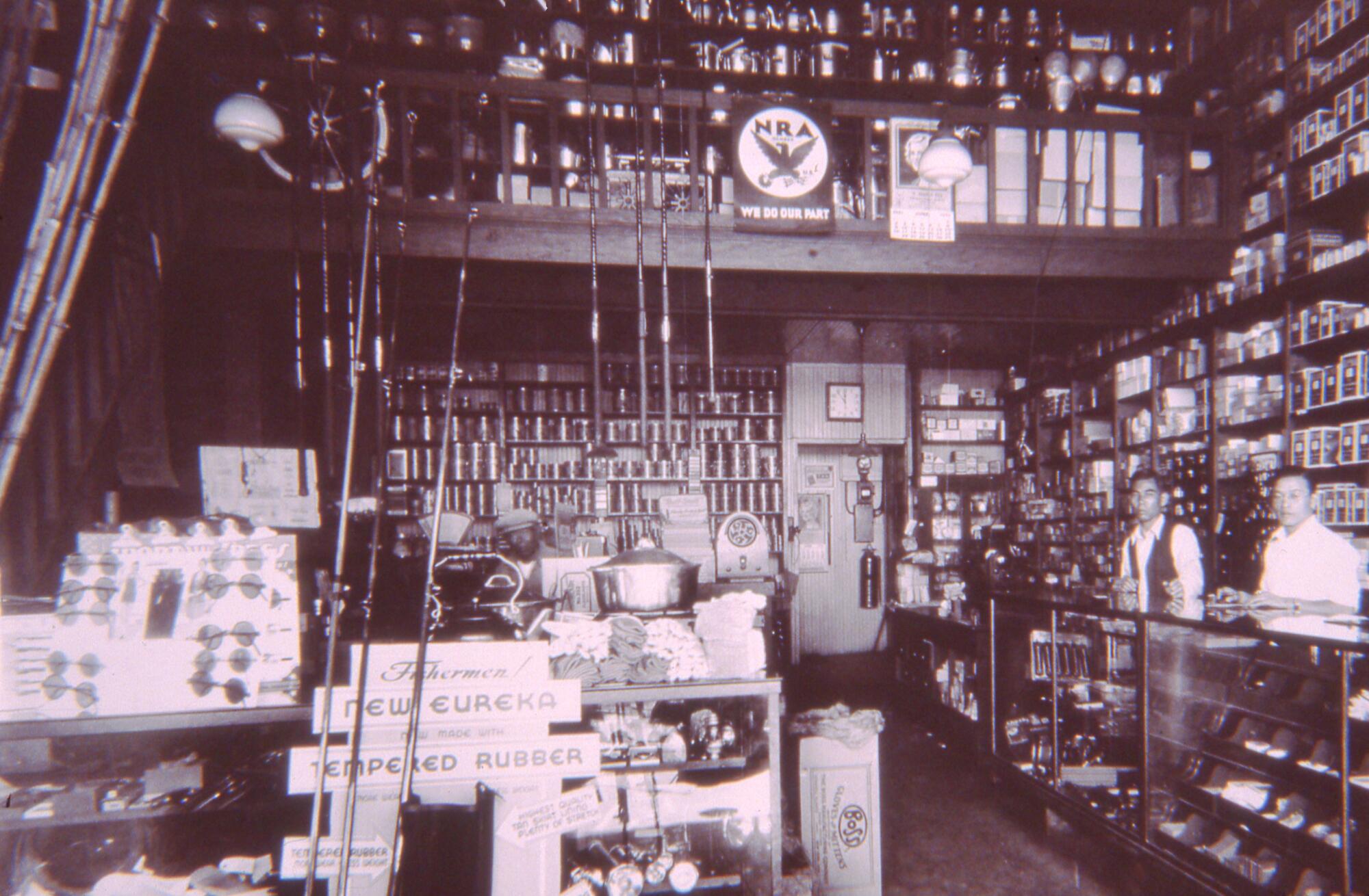 A vintage photo shows two men standing behind the counter of a store, its walls lined with fishing poles and other goods. 