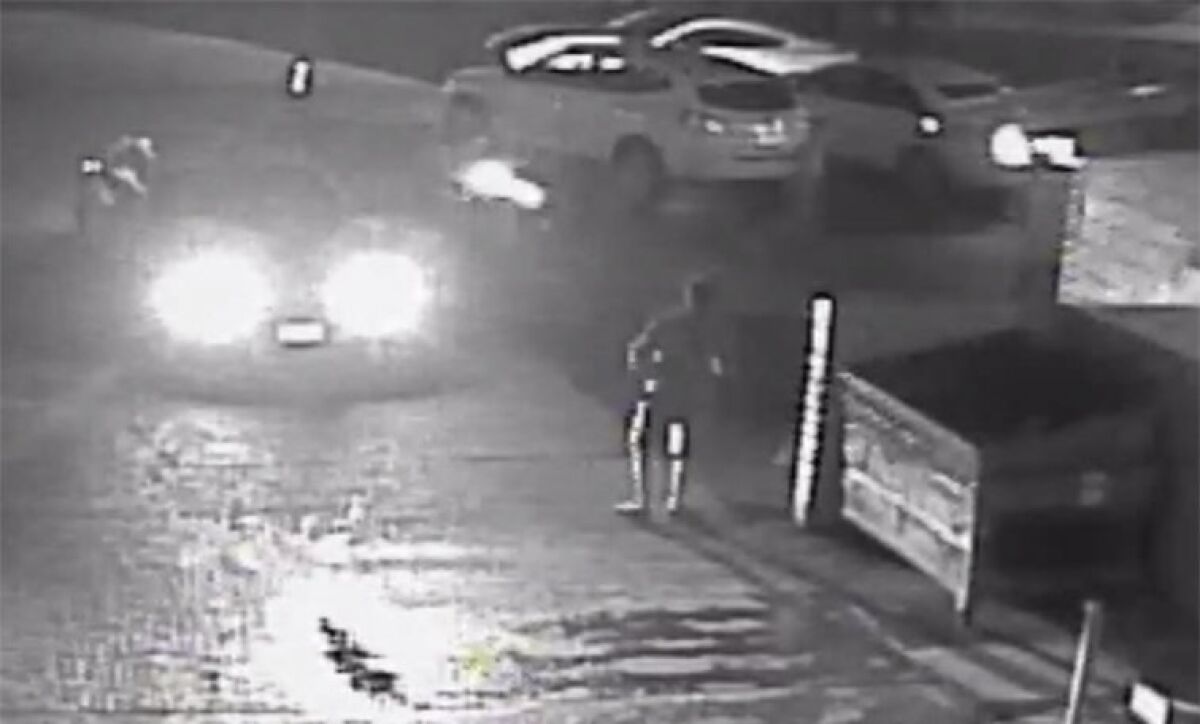 A still frame from surveillance video shows a man near a dumpster and a car shining its lights on him
