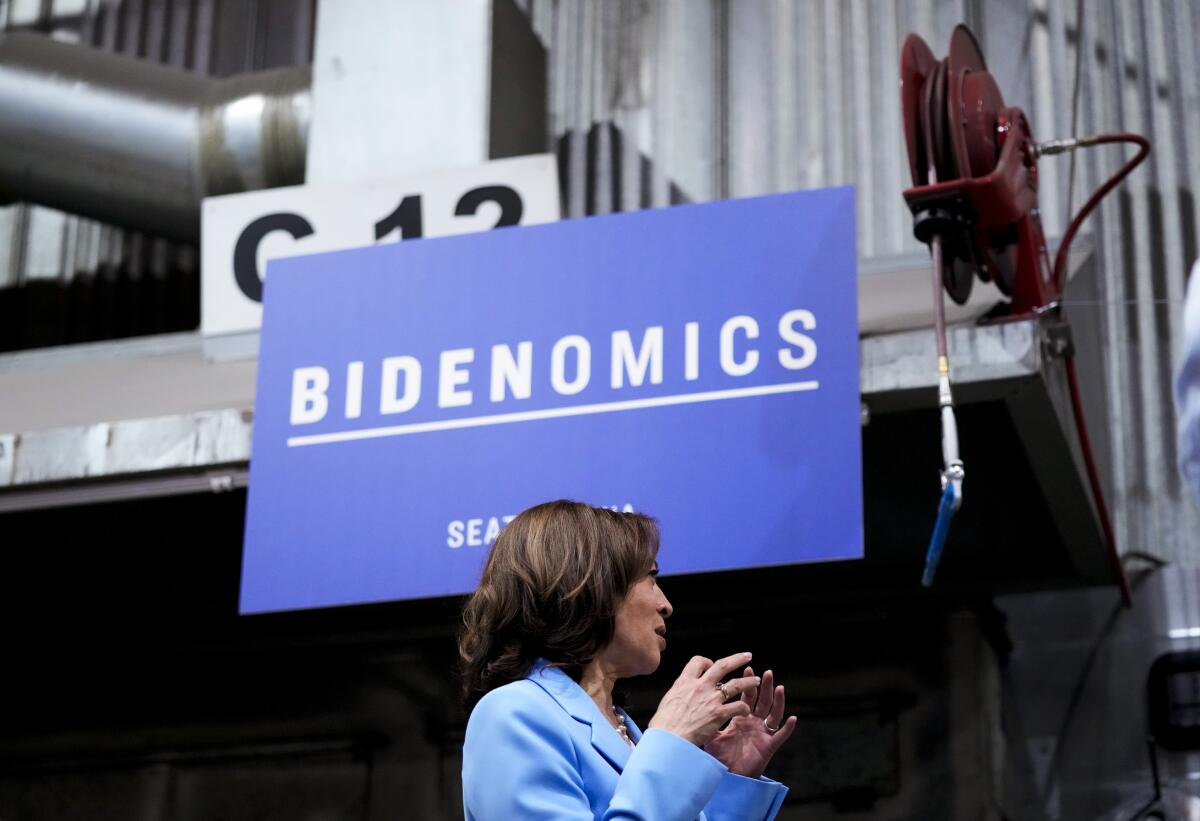 Vice President Kamala Harris gesturing with both hands, is silhouetted against a blue sign reading "Bidenomics" 