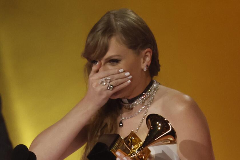 Taylor Swift holds her hand over her face in surprise and holds a Grammy in her other hand 