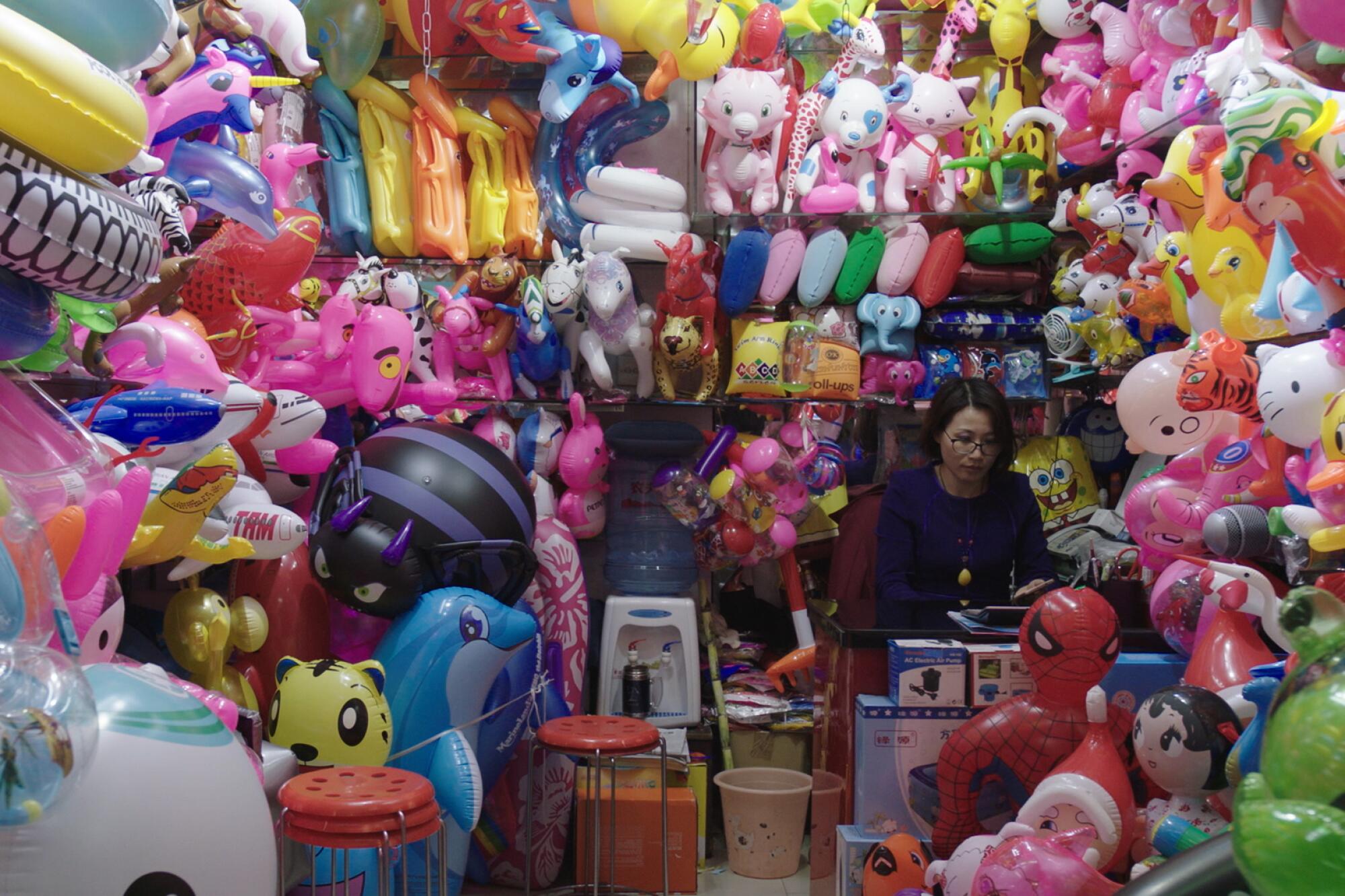 A video still shows an Asian woman sitting in a booth filled with plastic inflatable toys.