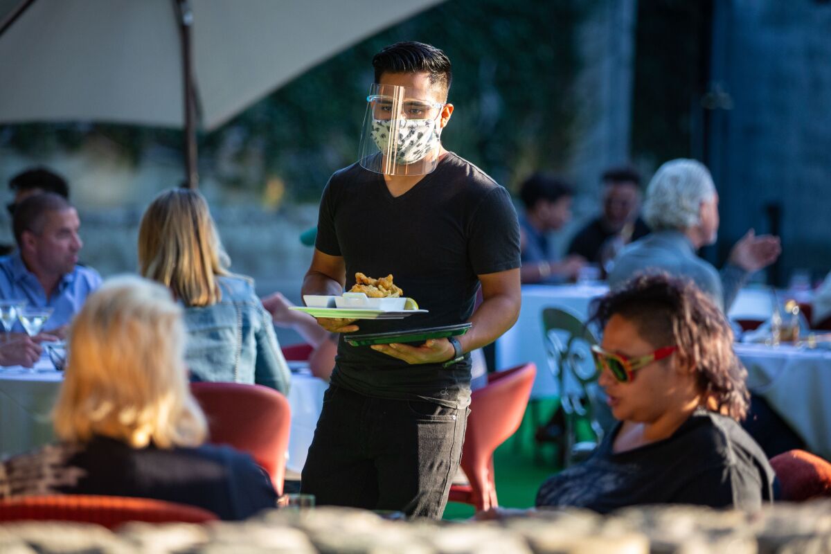 A server in a mask and face shield carries food to a table of diners at an outdoor seating area