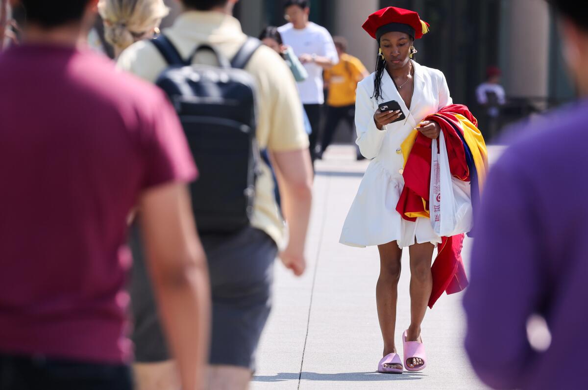 A woman in a white dress and pink slides holds graduation robes over one arm as she walks on a sidewalk.