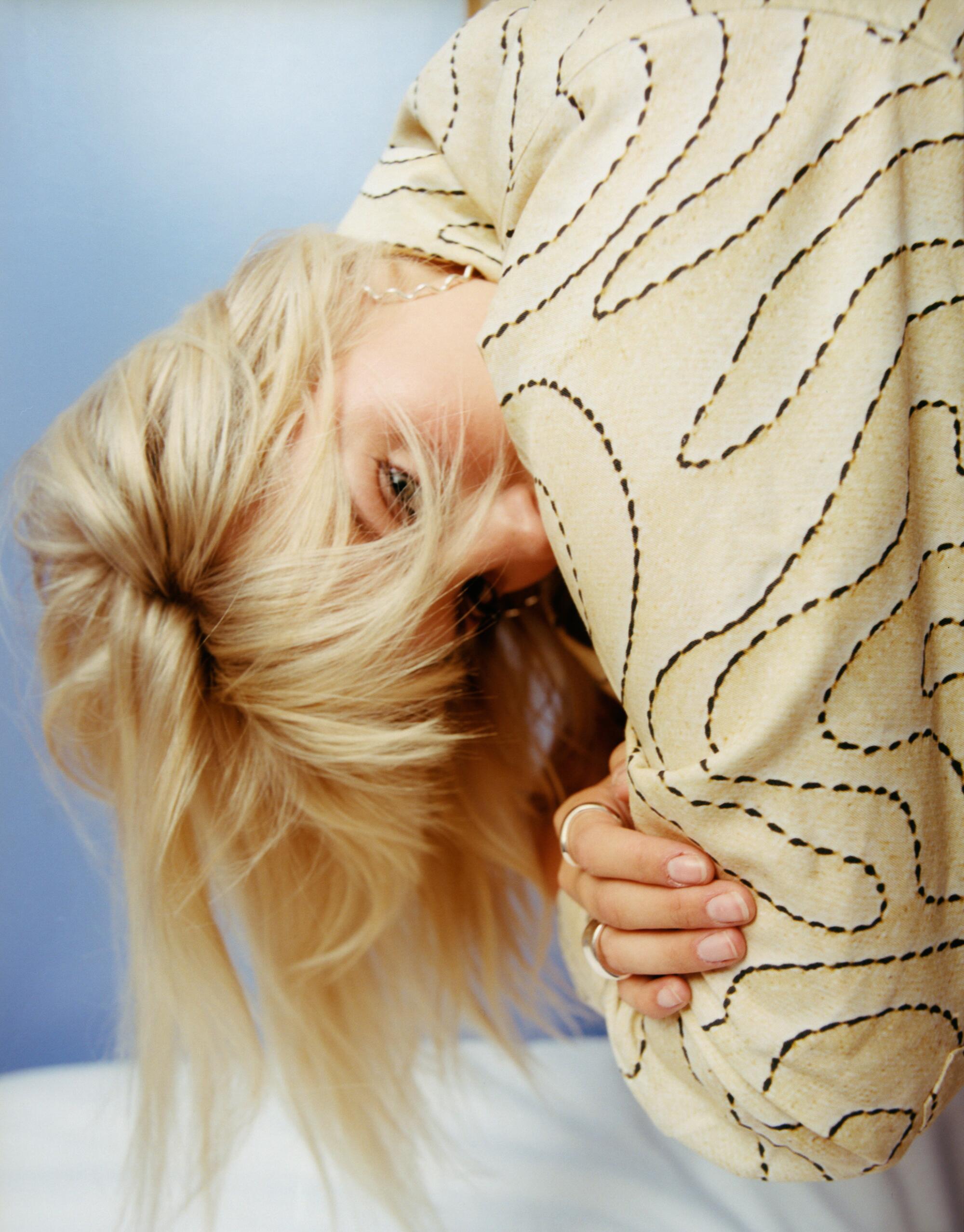 Reneé Rapp bends over with her head down as blond hair obscures her face and a patterned shirt hides her mouth