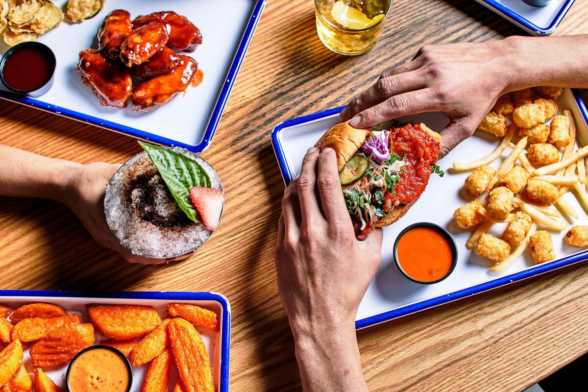 An assortment of foods on white rectangular plates with blue rims and hands holding a stuffed sandwich.