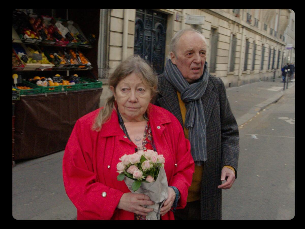 An older couple stands on the street outside a building, the woman holding flowers wrapped in newspaper.