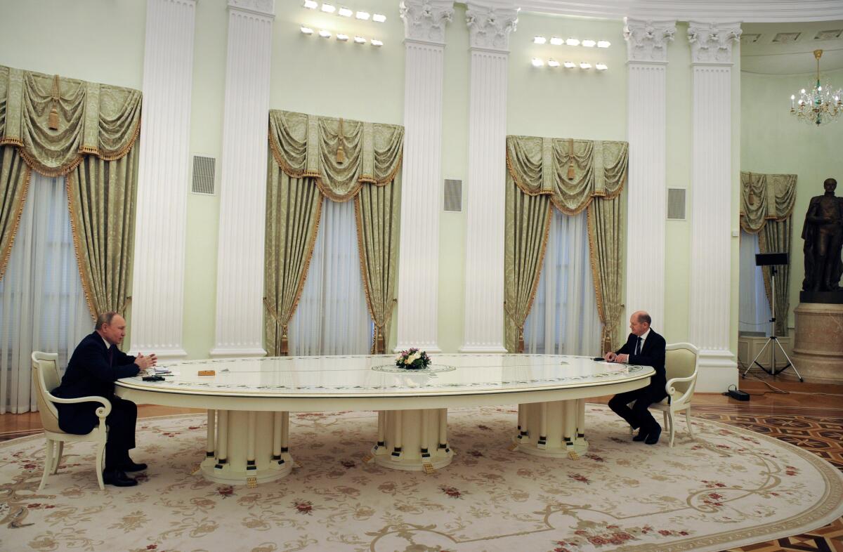 Two men in dark suits are seated at the opposite ends of a long cream-colored table in an ornate room