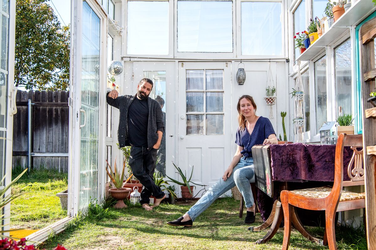 Trevor Morris stands in the greenhouse and Jenny Grosso sits at the greenhouse's dining table. 