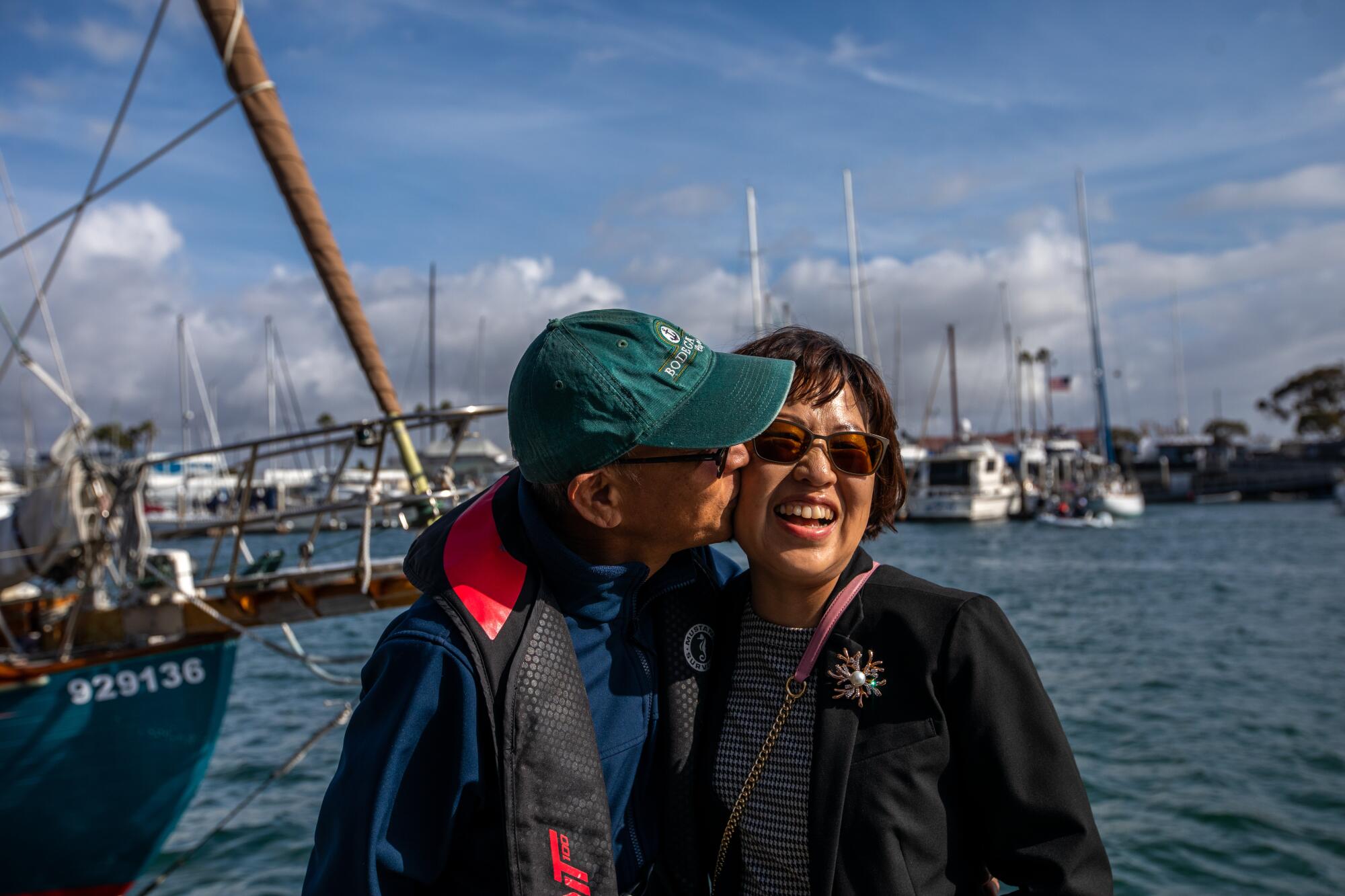 Standing in a sailboat harbor, a man at left kisses a woman who reacts smiling and closing her eyes.
