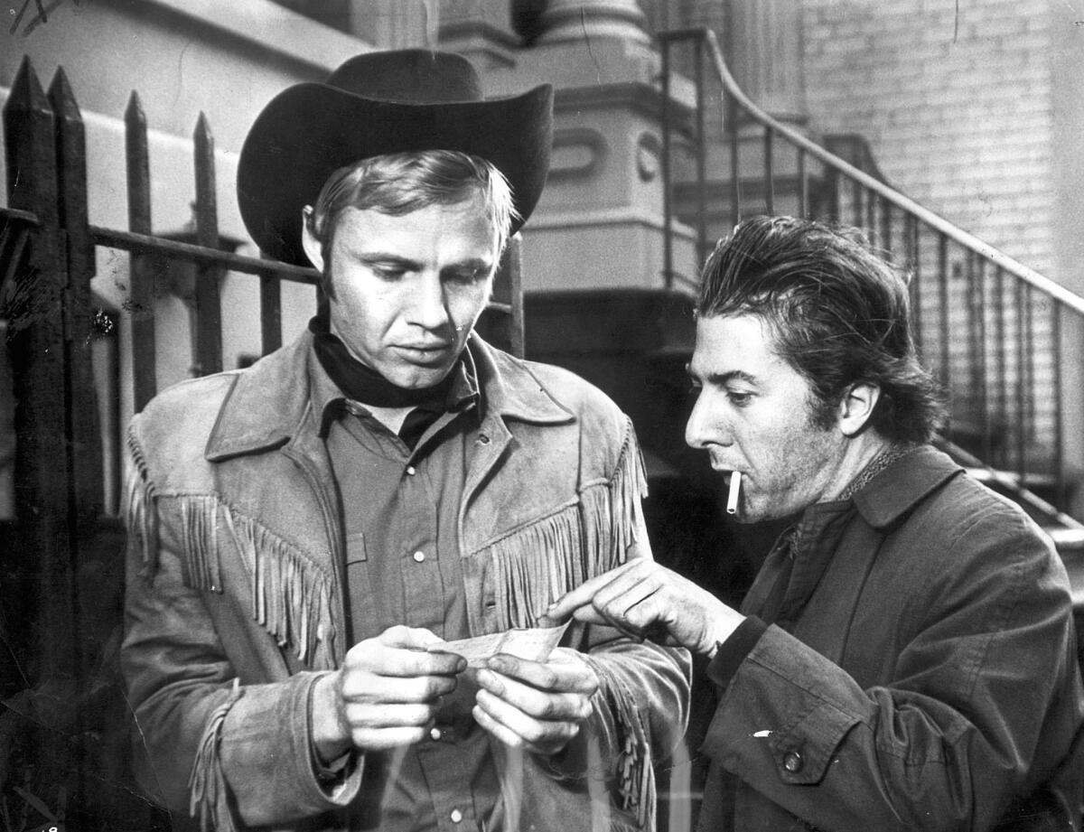 A man in a cowboy hat and fringed jacket and a man with a cigarette hanging from his mouth stand on a New York street.