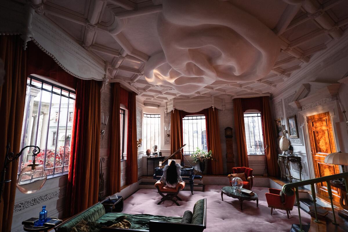 A woman sits at a piano in a room with a ceiling that is curved and creviced in a scene from "Poor Things"