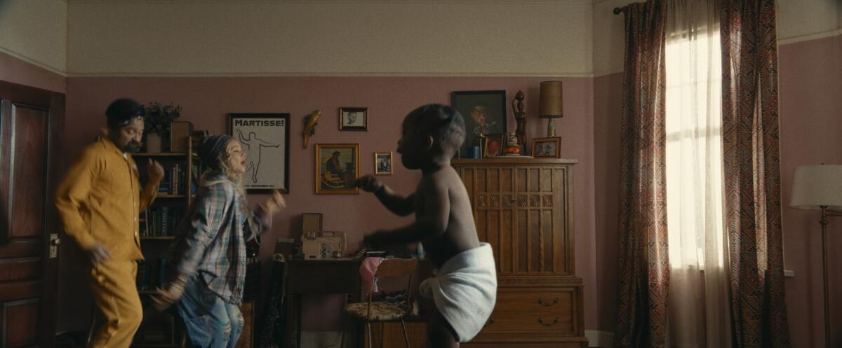 A man and a woman of normal stature dance next to a giant baby in a pink living room.