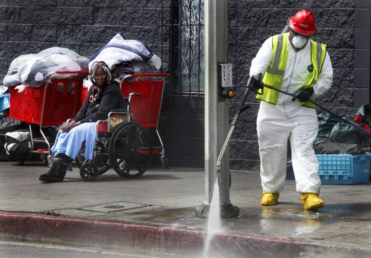 A homeless woman named Patricia watches from her wheelchair near the corner of 6th Street and Gladys Avenue while a sanitation worker uses a pressure washer to clean the streets of skid row.