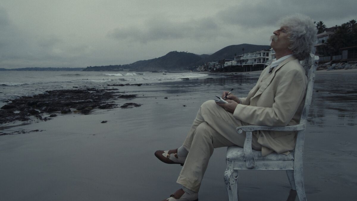 Val Kilmer dressed as Mark Twain sits in a weathered chair on a rocky beach.