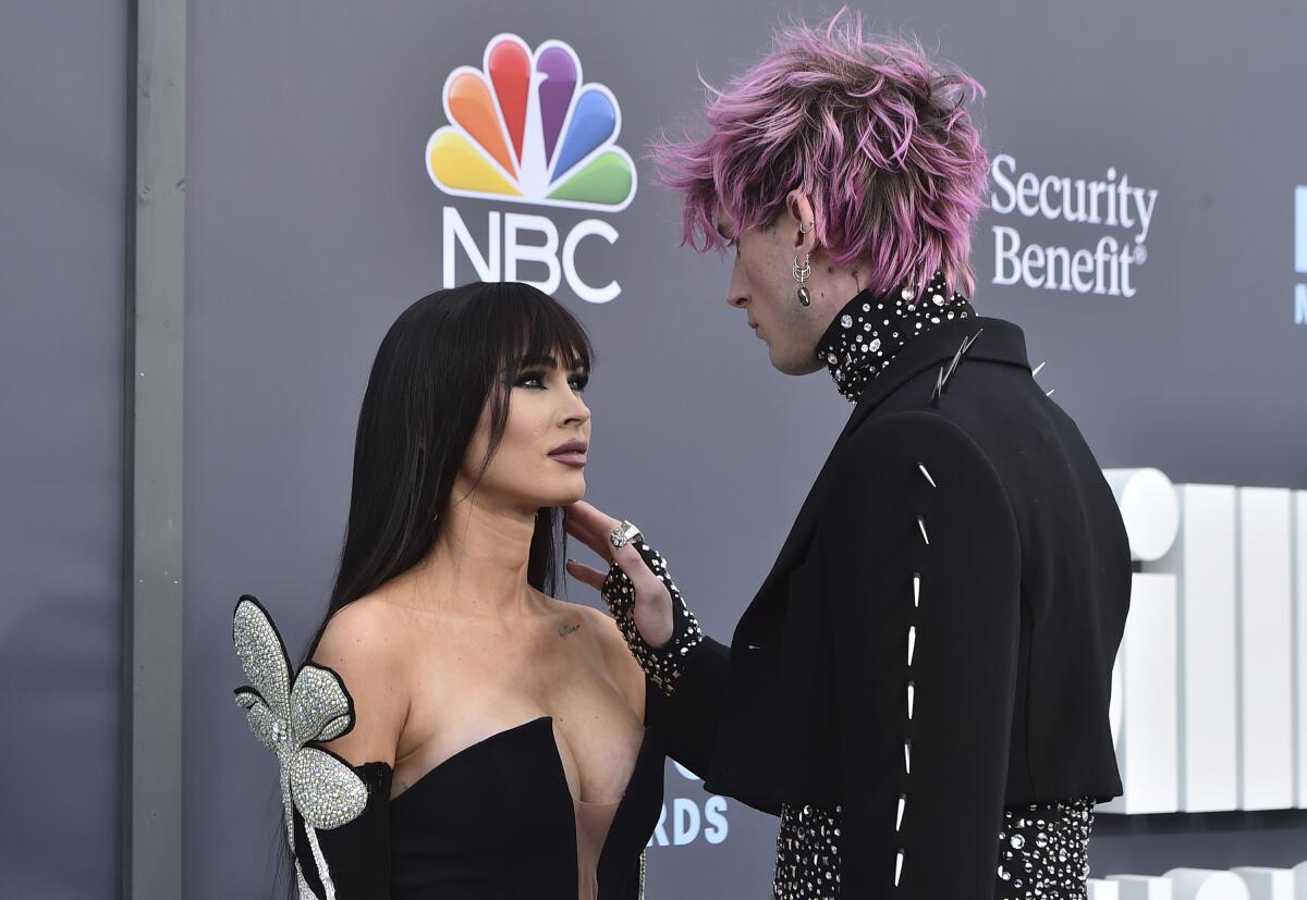 A woman with long dark hair looks up at her pink-haired male partner on an awards show red carpet