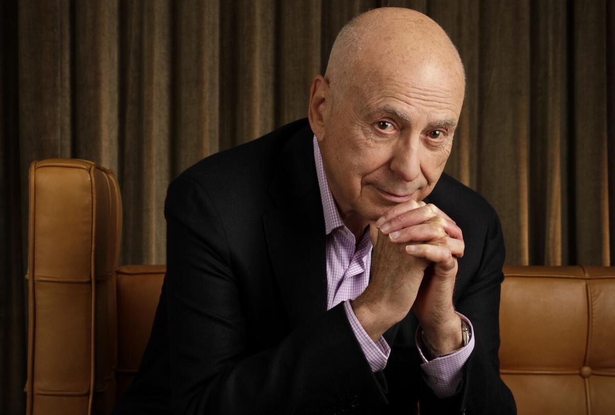 Alan Arkin leans forward from his seat with his hands folded underneath his chin.