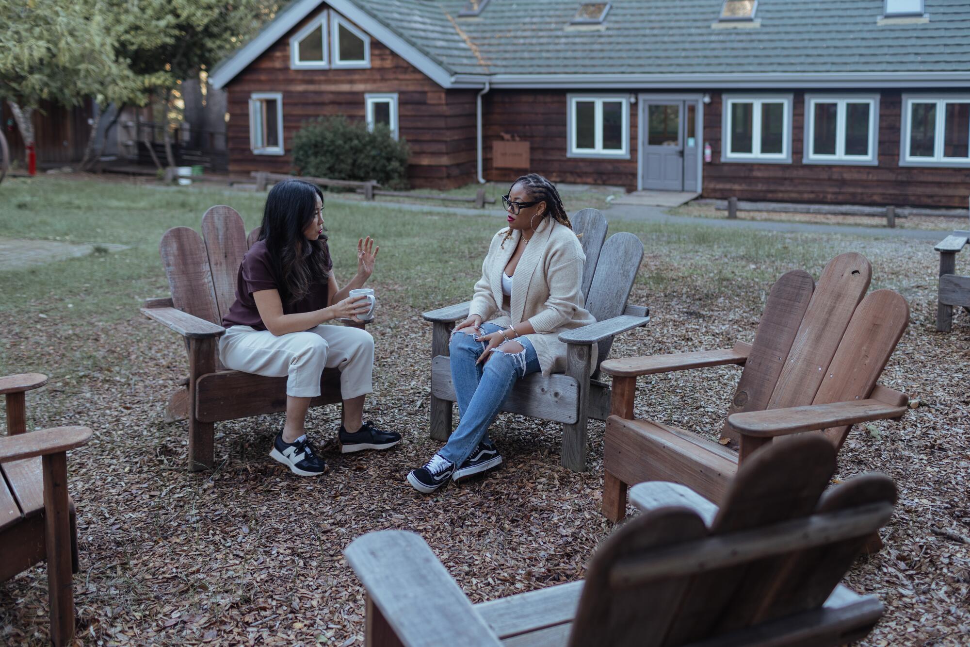 Two women sit in Adirondack chairs, in a circle with three other empty ones 