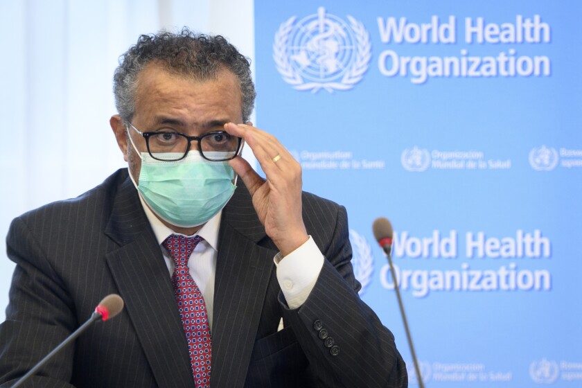 Tedros Adhanom Ghebreyesus, in a mask, straightens his glasses with one hand as he sits behind two microphones.