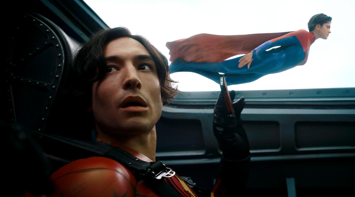 Ezra Miller stares with their mouth agape inside an aircraft as a woman in a superhero costume flies outside the the window.