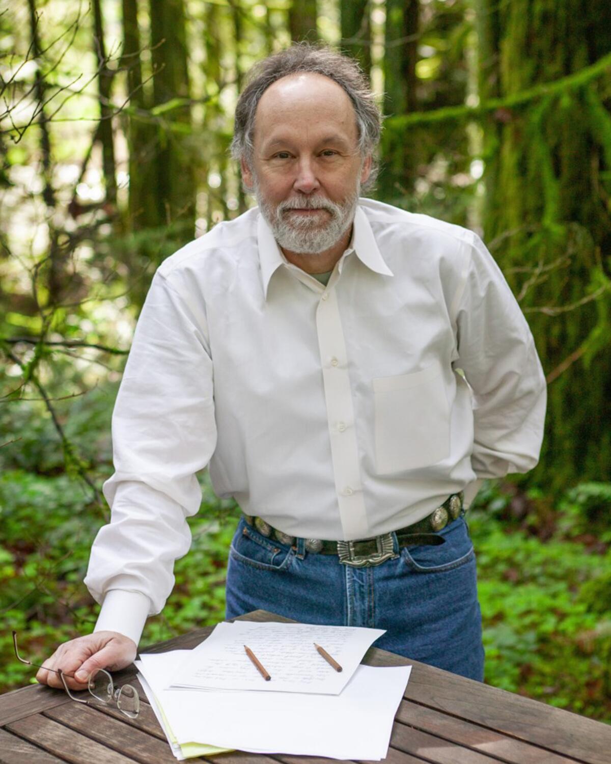 Barry Lopez stands in a forested spot with one hand on a wood table that has papers and two pencils.