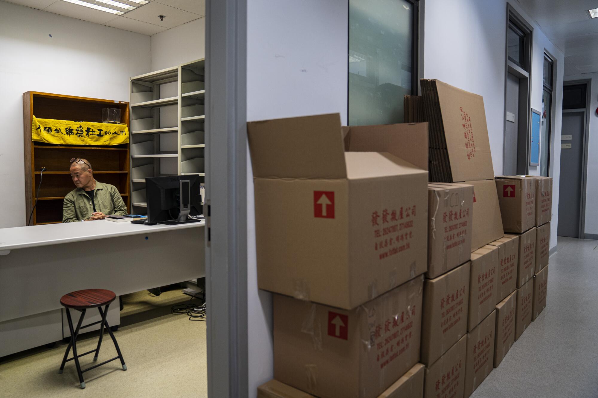 A man sits at a desk in an empty office with cardboard moving boxes piled next to the wall outside.