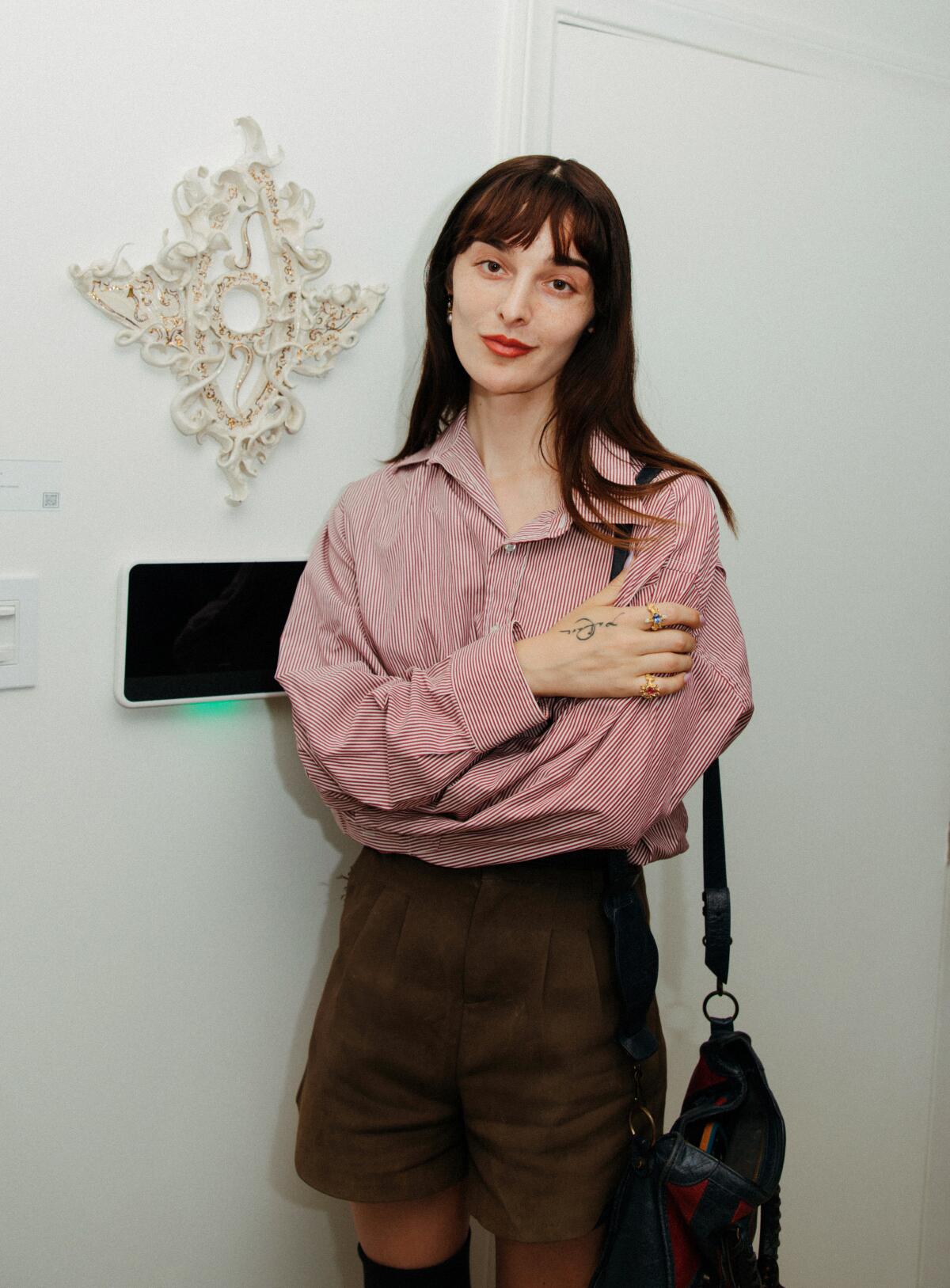 An artist with dark hair, wearing a red top and brown shorts, stands in front of her art displayed on a white wall.