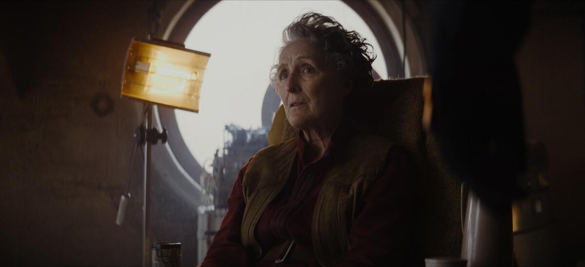 A older woman sitting before a porthole window next to a lamp.
