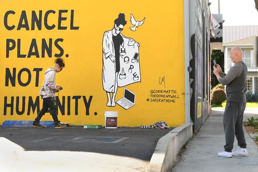 WEST HOLLYWOOD, CALIFORNIA MARCH 25, 2020-Artist Corie Mattie paints a mural that says :"Cancel Plans Not Humaity" on the side of a pop-up store as a man takes a picture in West Hollywood Wednesday. (Wally Skalij/Los Angeles Times)