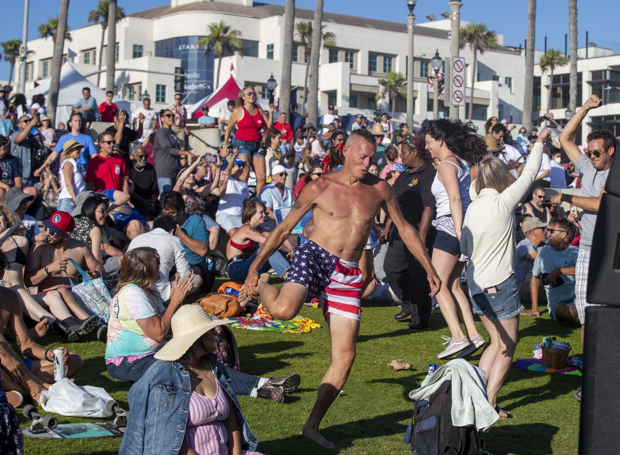 A man wearing shorts in U.S. flag colors holds his ankle with one hand near people dancing on a lawn
