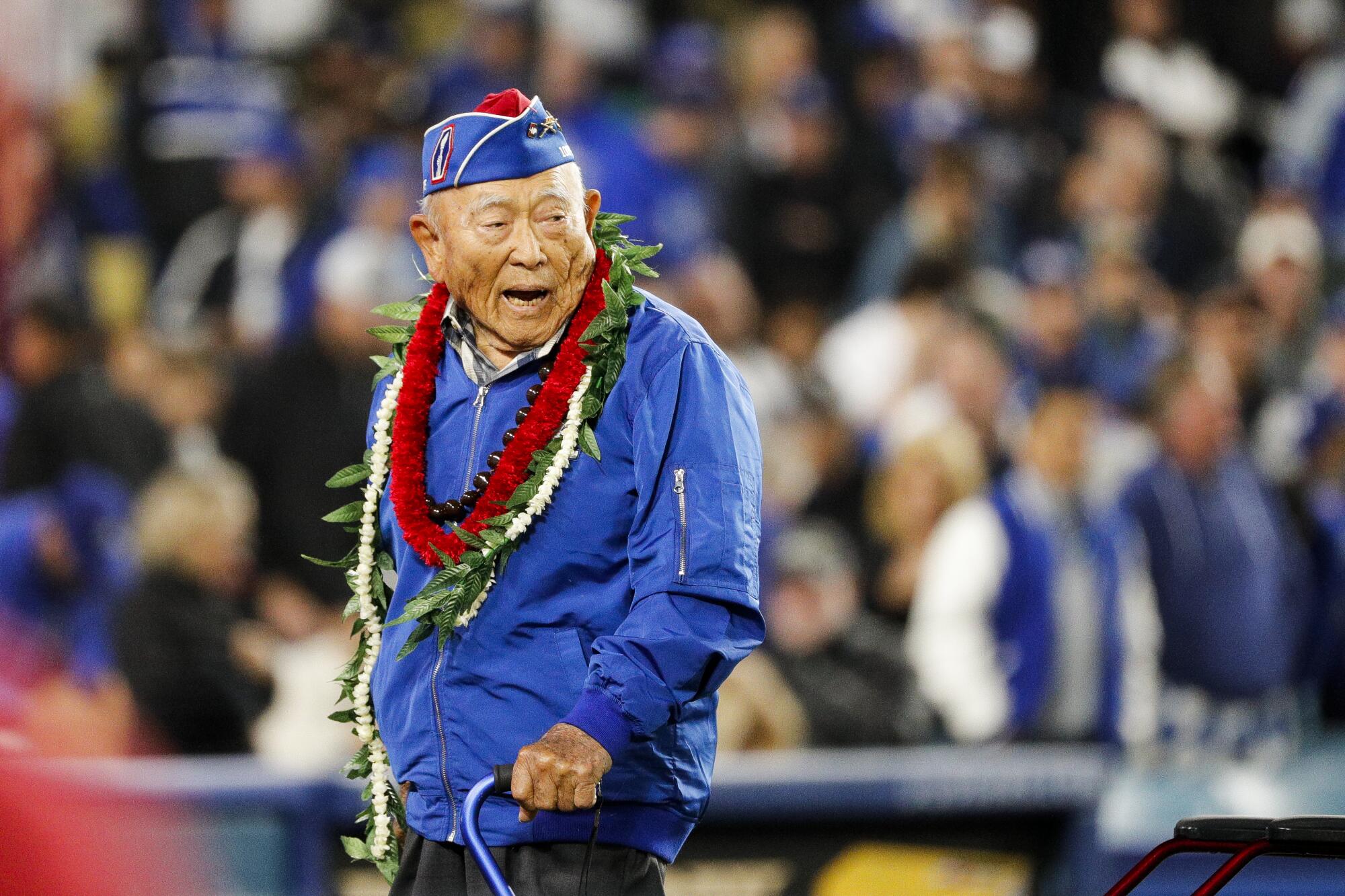 An elderly man wearing a windbreaker and military cap with leis around his neck and carrying a cane smiles