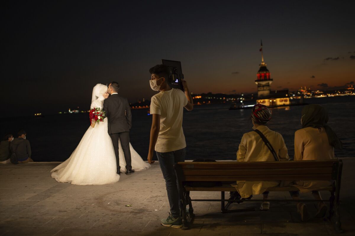 A wedding photographer's assistant, wearing a mask for protection against the spread of coronavirus, holds lighting equipment, as a couple poses for photographs backdropped by Istanbul's iconic Maiden's Tower (Kiz Kulesi) at the Bosphorus Strait separating the European and Asian sides of Istanbul, Friday, Sept. 11, 2020. Turkey is getting tough on people who flout self-isolation rules despite testing positive for the coronavirus. An Interior Ministry circular sent to the country's 81 provinces on Friday said people caught leaving their homes despite isolation orders will be quarantined and supervised at state-owned dormitories or hostels. (AP Photo/Yasin Akgul)