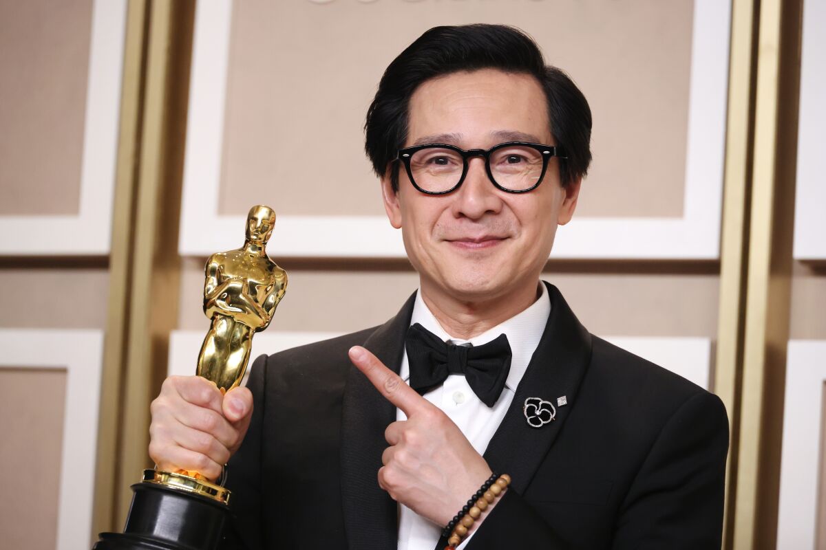 A man with short black hair wearing a tuxedo points at an Oscar trophy in his other hand