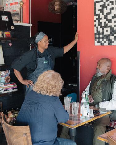 A woman chef stands talking to a seated couple in her restaurant