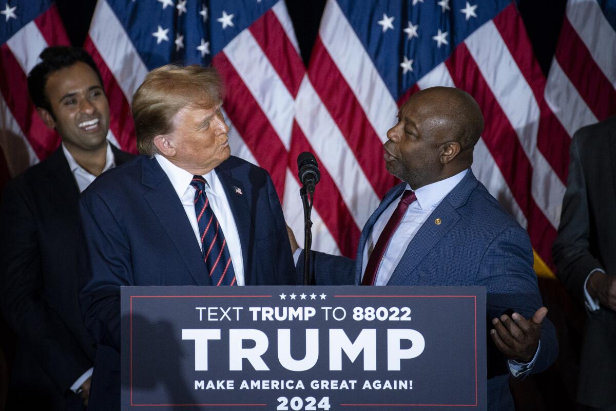 Donald Trump and Tim Scott stand at a lectern with a sign promoting Trump's 2024 presidential campaign