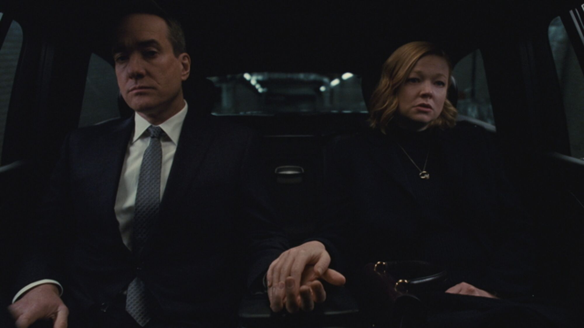 A husband and wife, dressed all in black, hold hands in the back of a car.