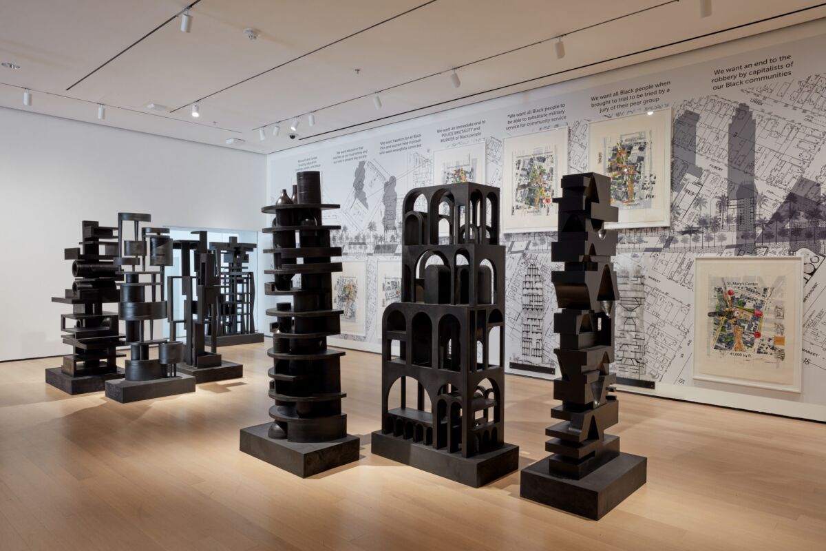 A group of vertical architectonic sculptures, all painted black, are seen in a museum gallery