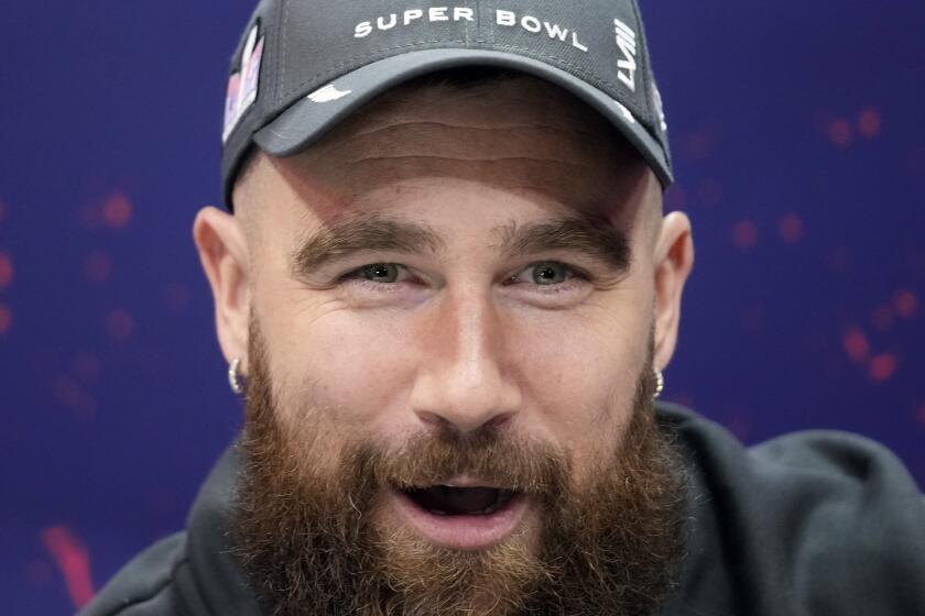 A close-up of a bearded Travis Kelce wearing a baseball cap and small hoop earrings