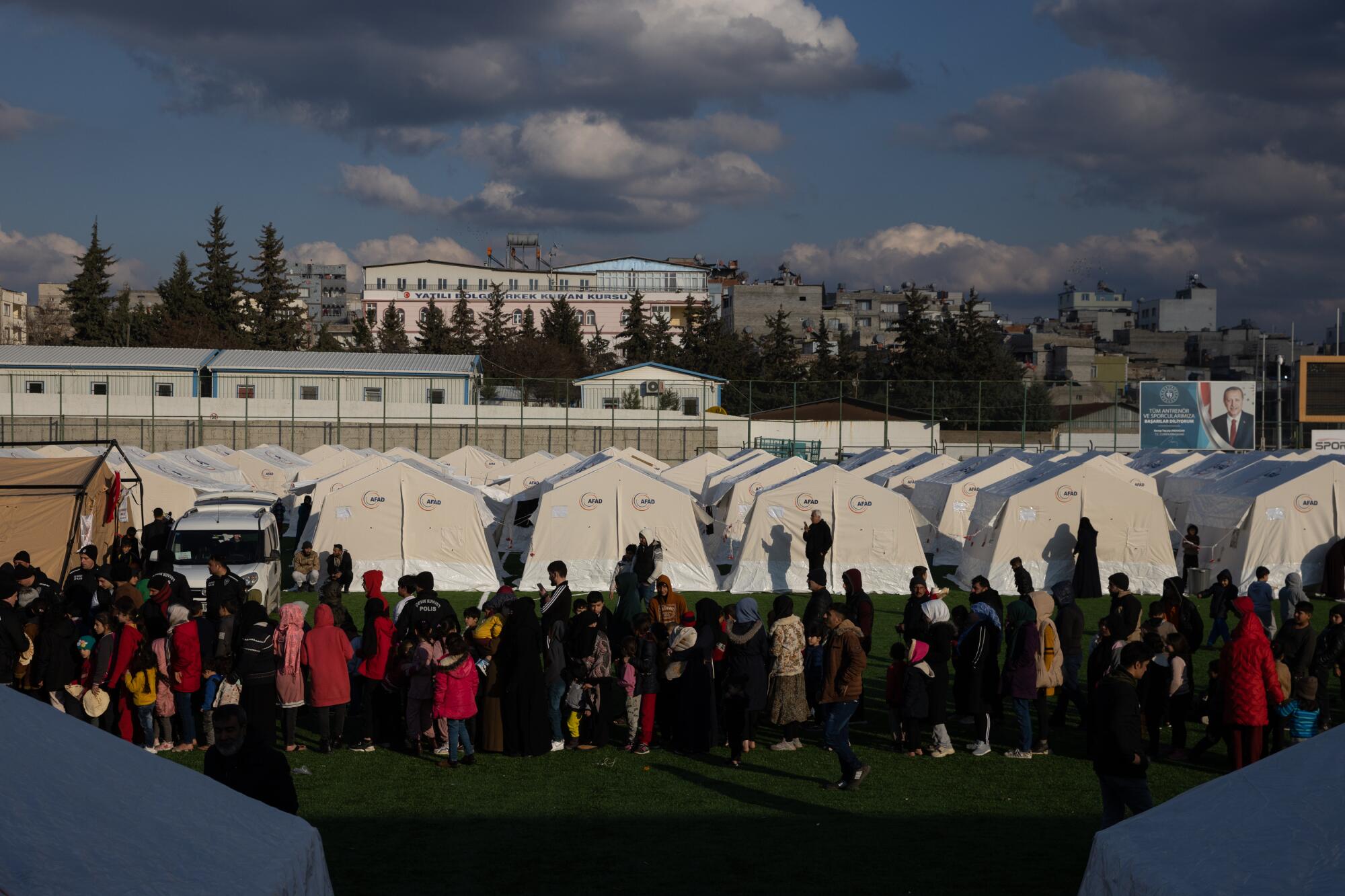 Displaced Syrians stand in line near a row of white tents.
