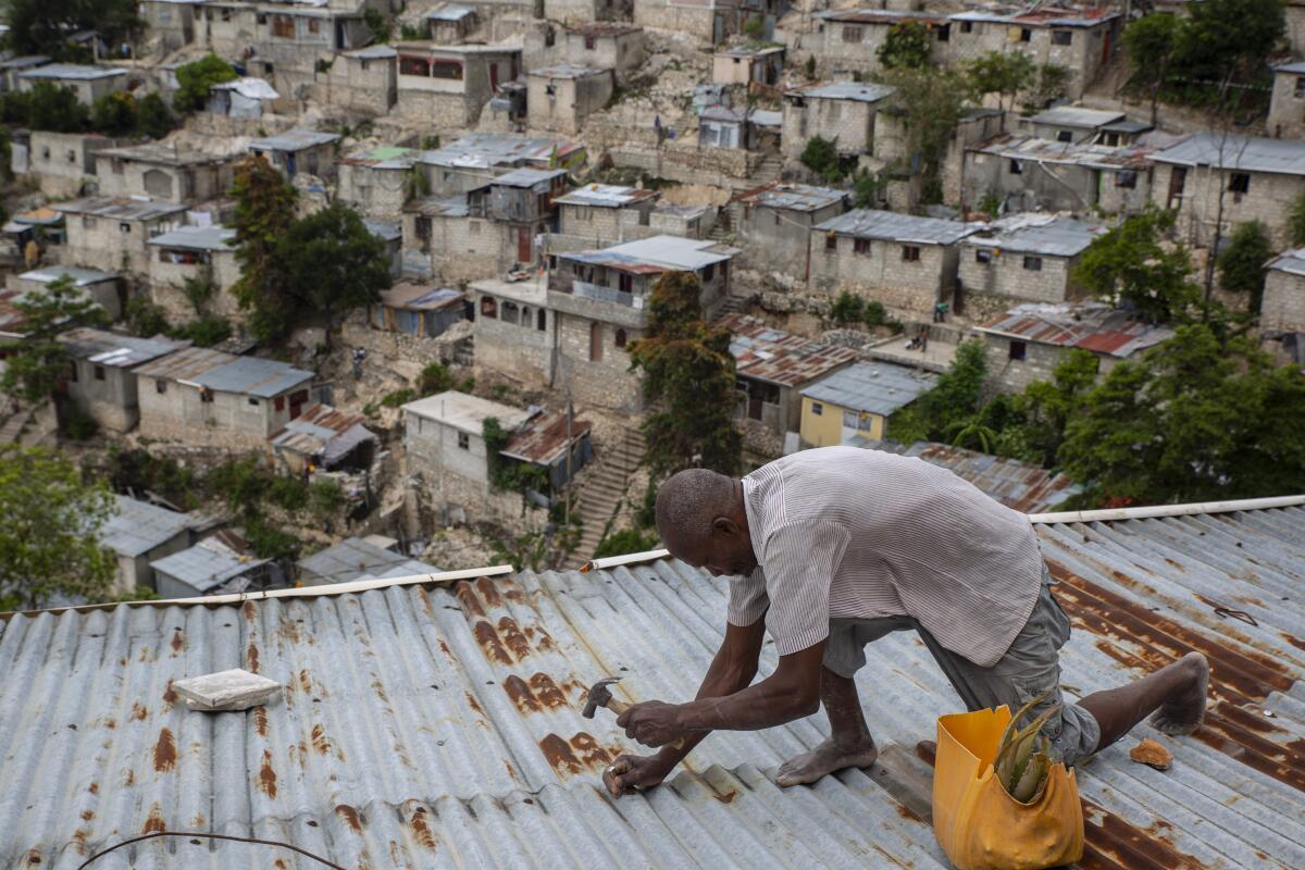 A man bends on one knee as he hammers a nail into a corrugated metal roof.