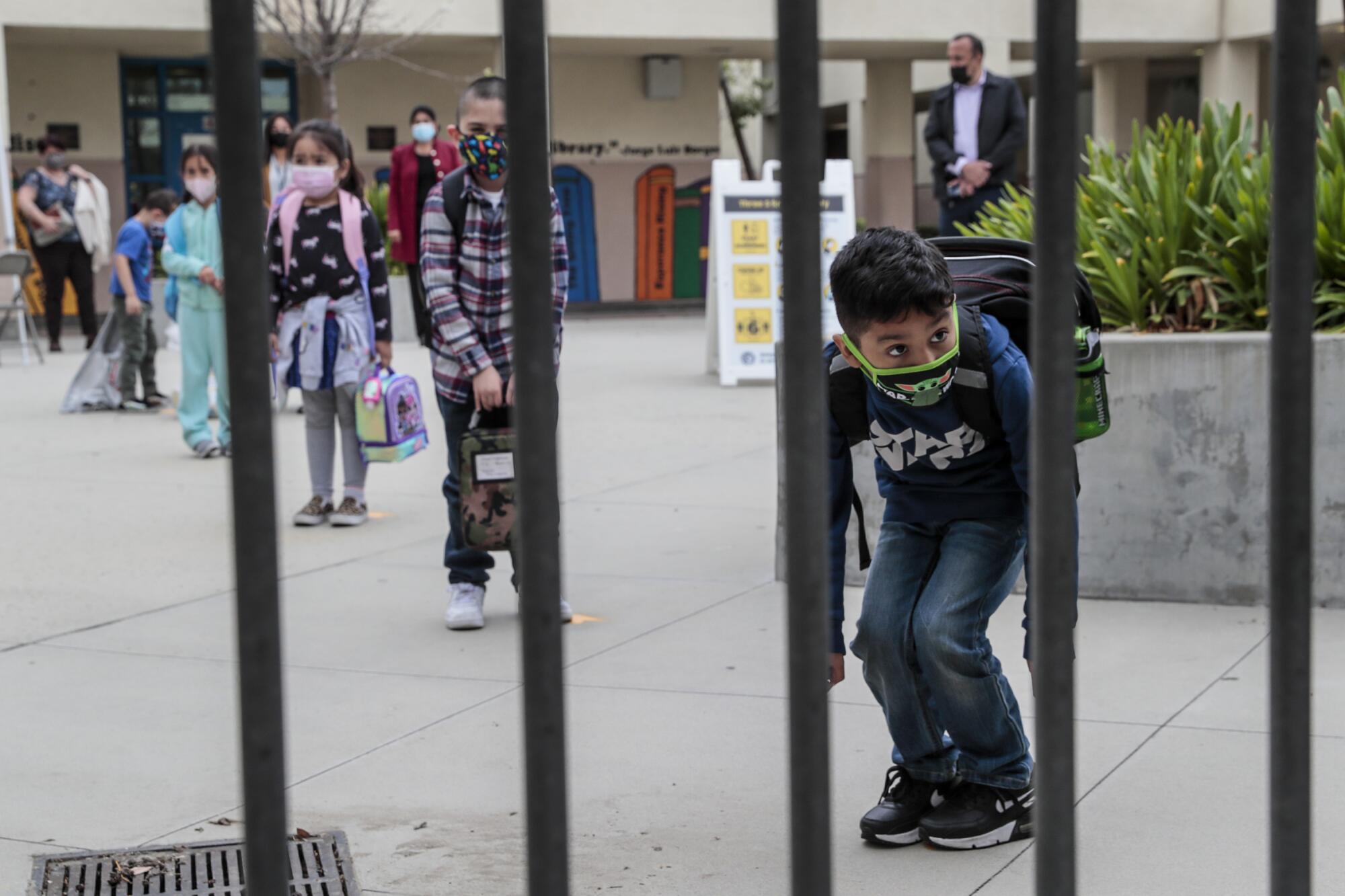 A boy in a mask and backpack bends and peeks through a gate.
