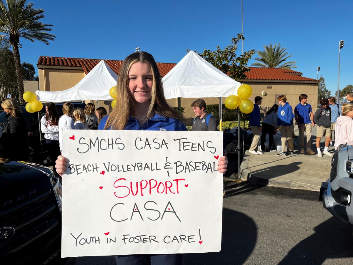 Grace Jackson holds a sign reading in part: "Beach Volleyball & Baseball Support CASA Youth in Foster Care!"
