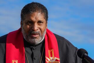  Rev. Dr. William Barber speaking in front of a microphone while wearing a black robe and red sash
