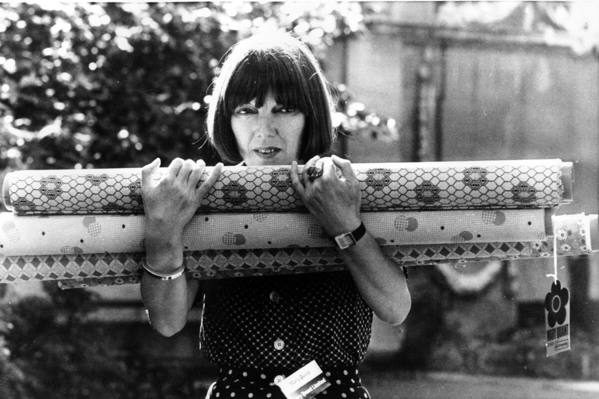A black and white photo from the '70s shows Mary Quant, in a page boy haircut, holding several bolts of fabric.