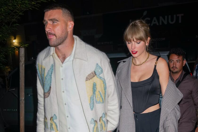 Travis Kelce is wearing a jacket and white shirt and is holding hands with Taylor Swift who is in a plaid coat